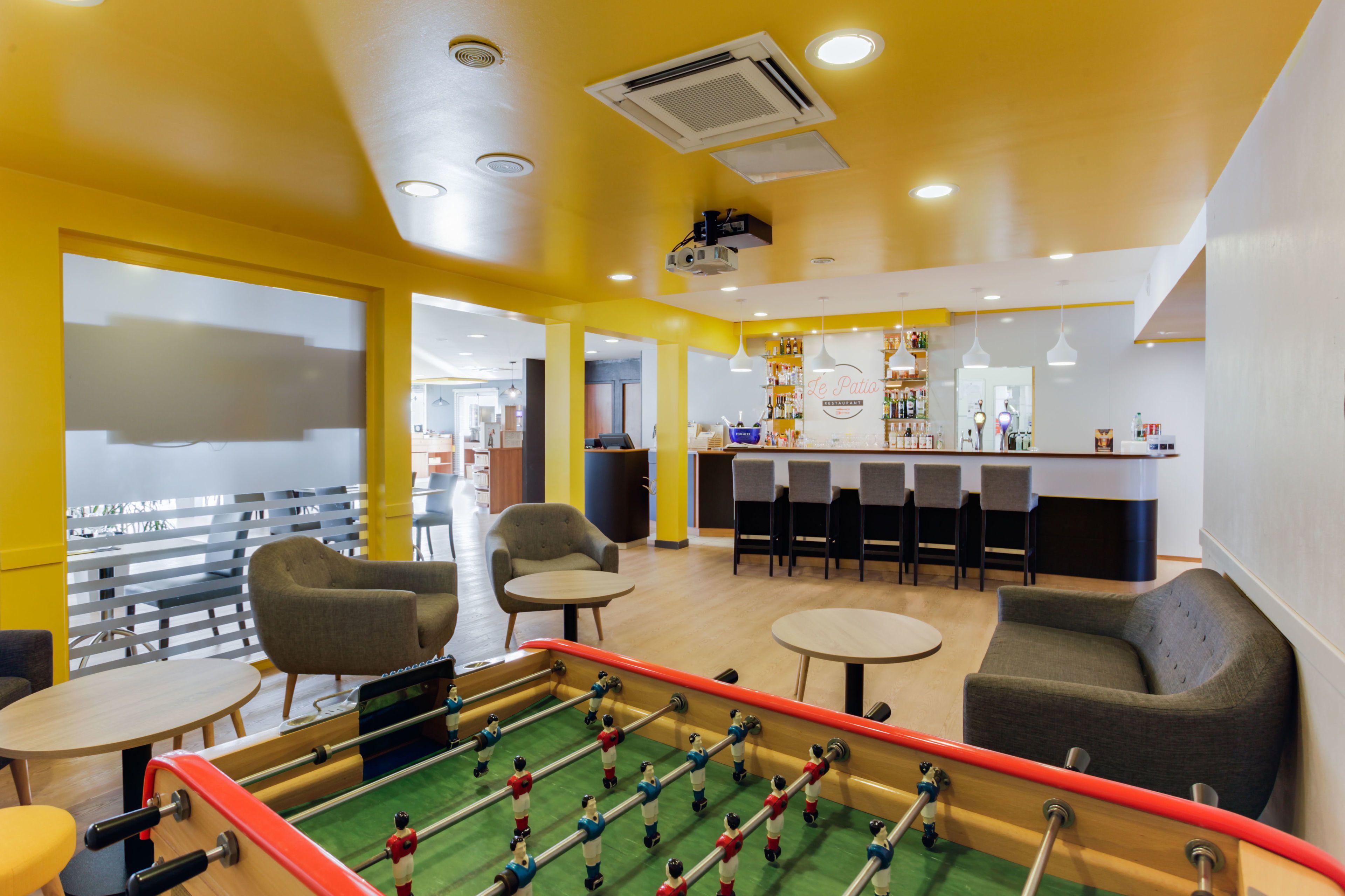 A brightly lit indoor space with a foosball table, seating areas, and a bar counter.