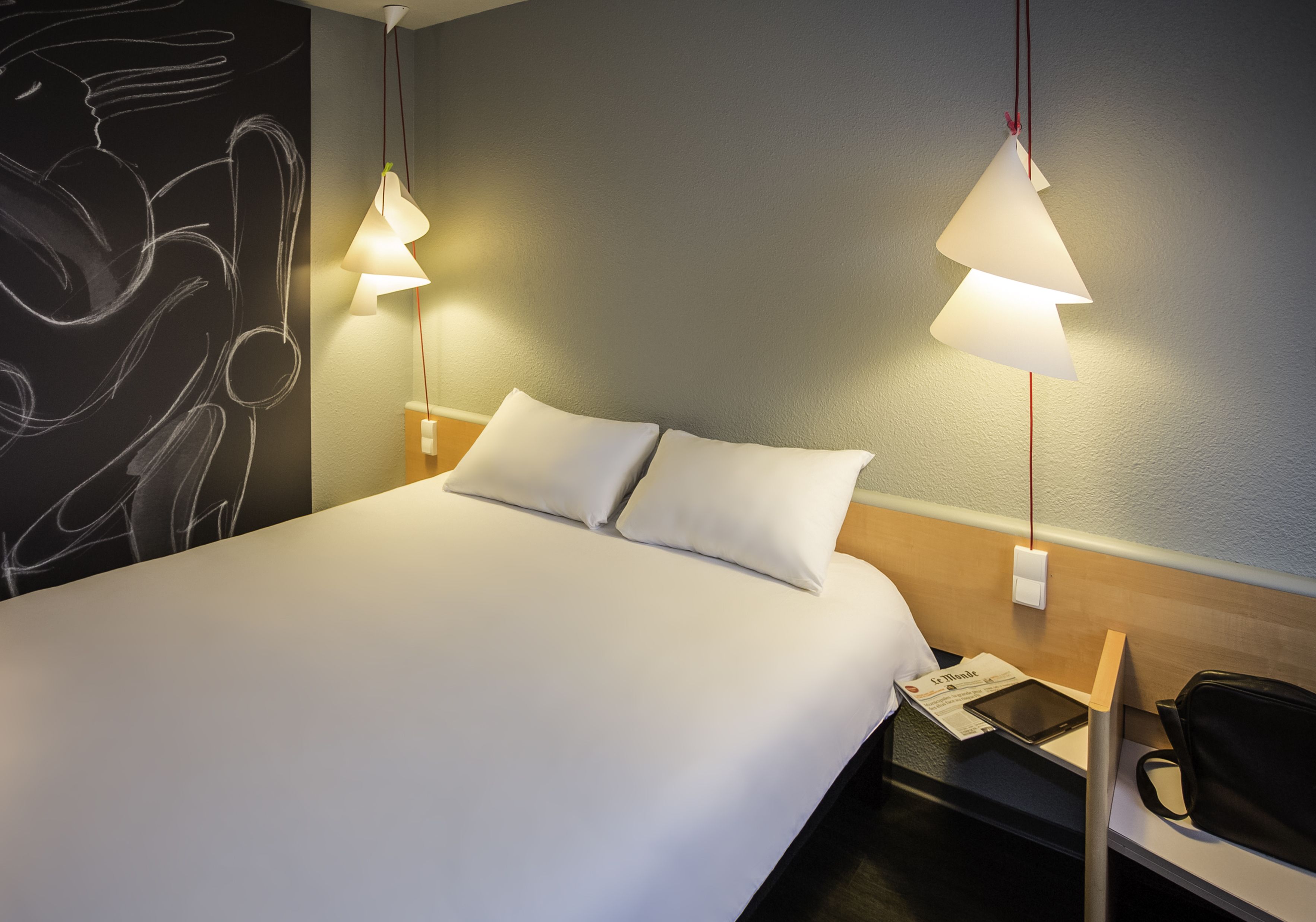 Une chambre d’hôtel avec un lit soigneusement fait, deux lampes de chevet, un mur orné d’œuvres d’art abstraites, une table de chevet avec une lampe, une chaise et un sac.