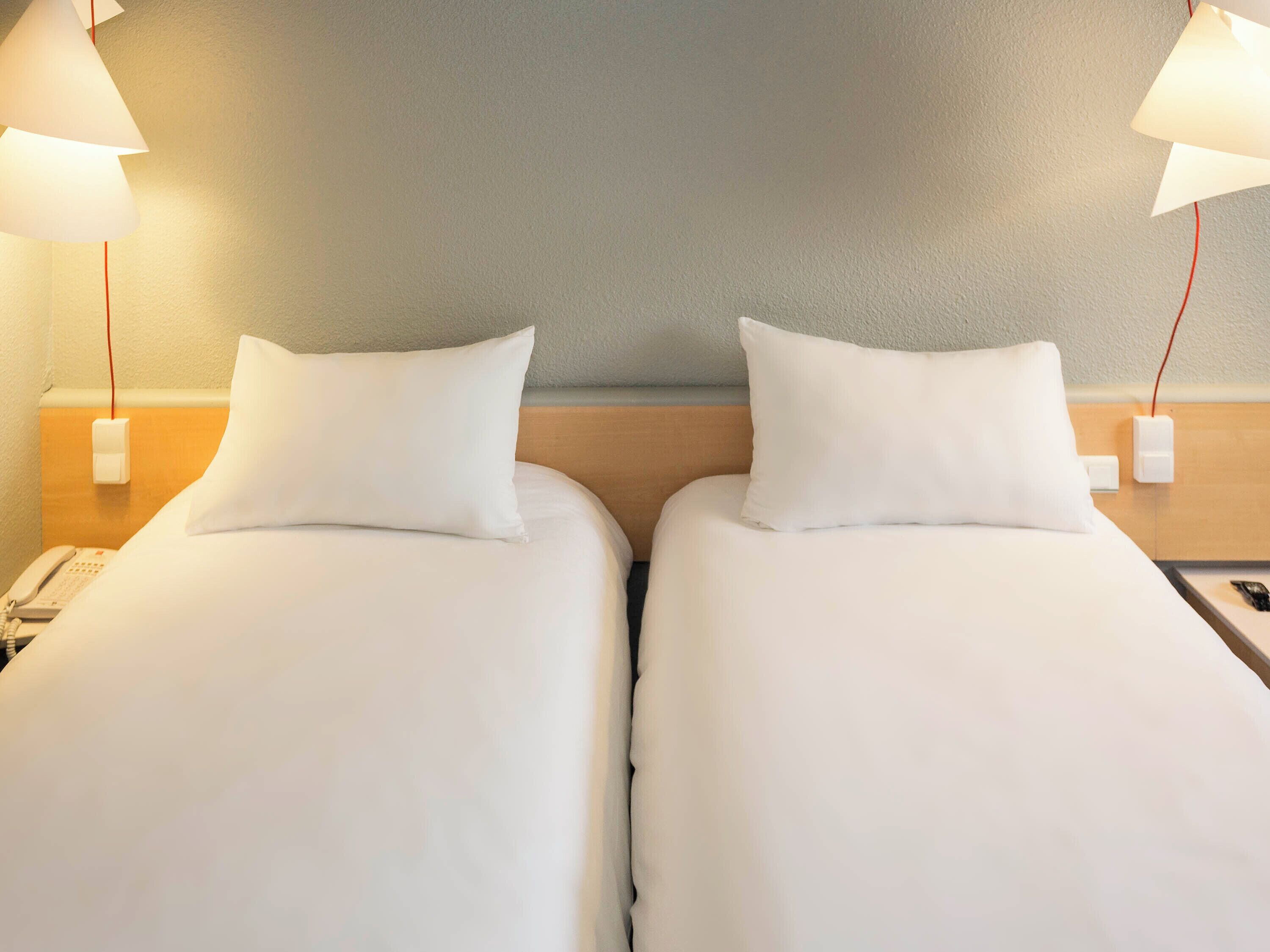 Une chambre d’hôtel avec deux lits simples, chacun équipé d’un oreiller et d’une table de chevet. Il y a un téléphone sur la table de chevet du lit de gauche et une télécommande sur celle du lit de droite.