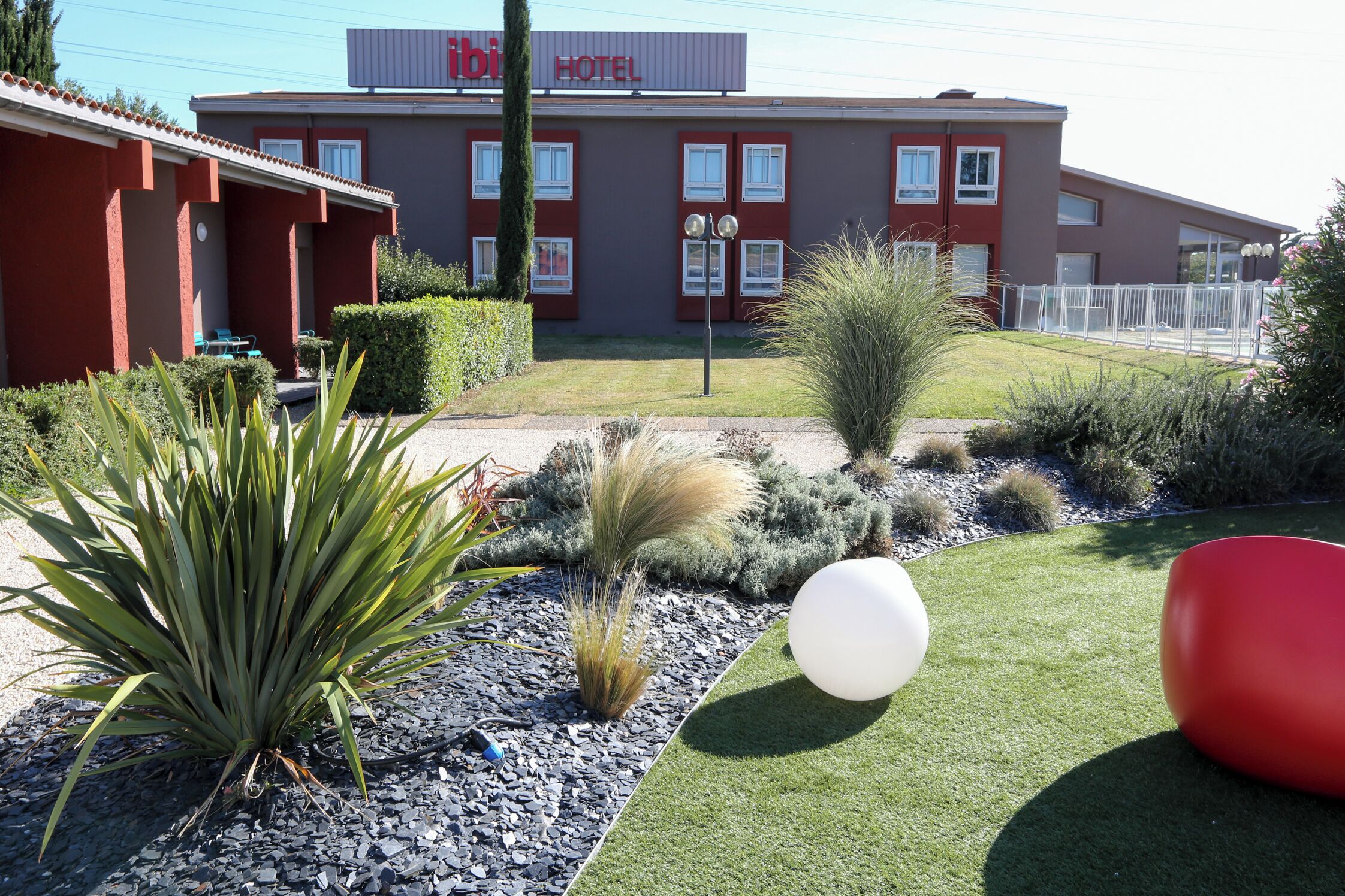 Un hotel con fachada roja, un cartel y un jardín bien cuidado con diversas plantas y un objeto esférico blanco.