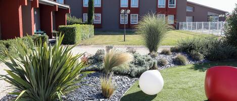 Un hotel con fachada roja, un cartel y un jardín bien cuidado con diversas plantas y un objeto esférico blanco.