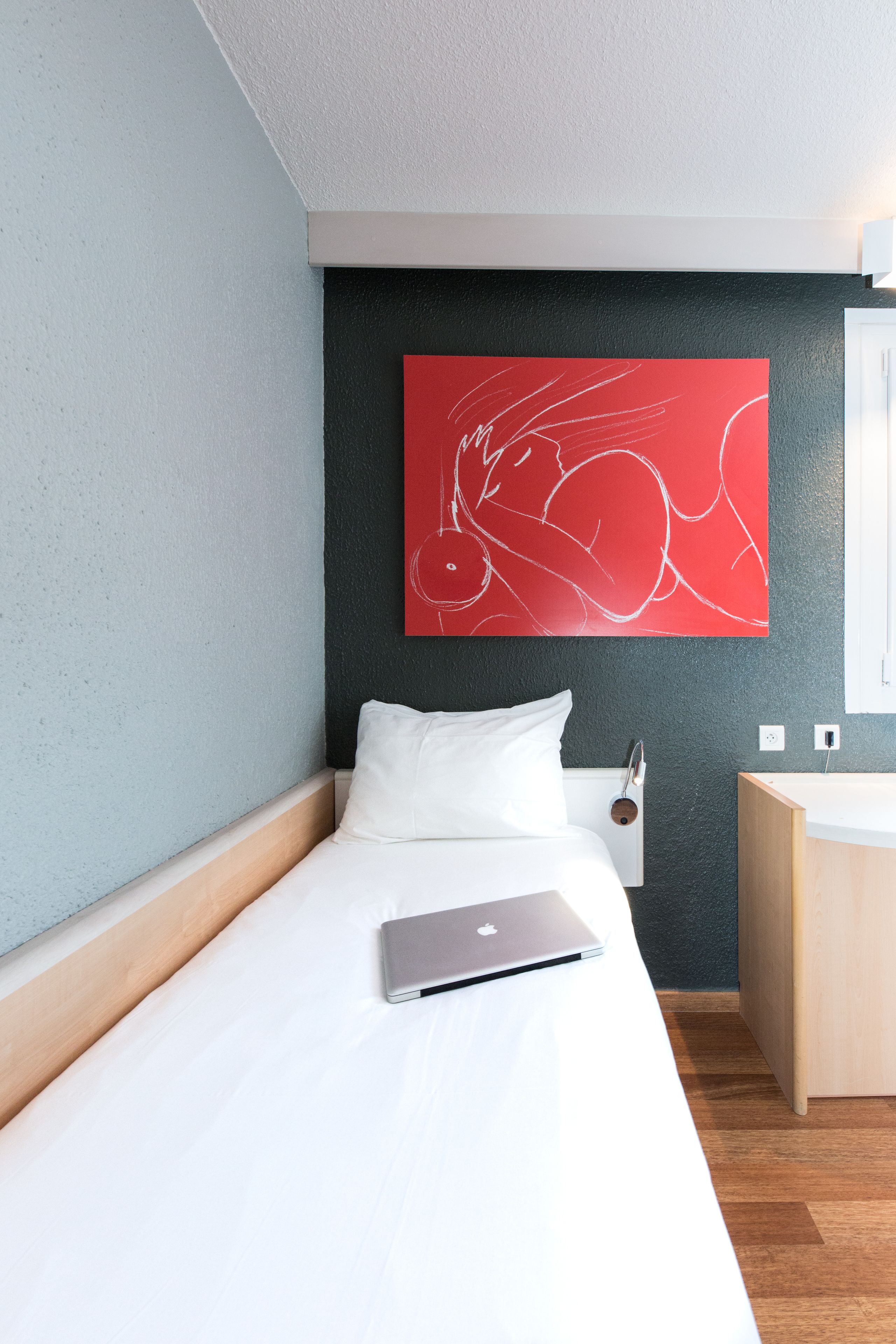 Une chambre d’hôtel moderne avec un lit, un ordinateur portable, une œuvre d’art abstraite rouge accrochée au mur et une table de chevet en bois.