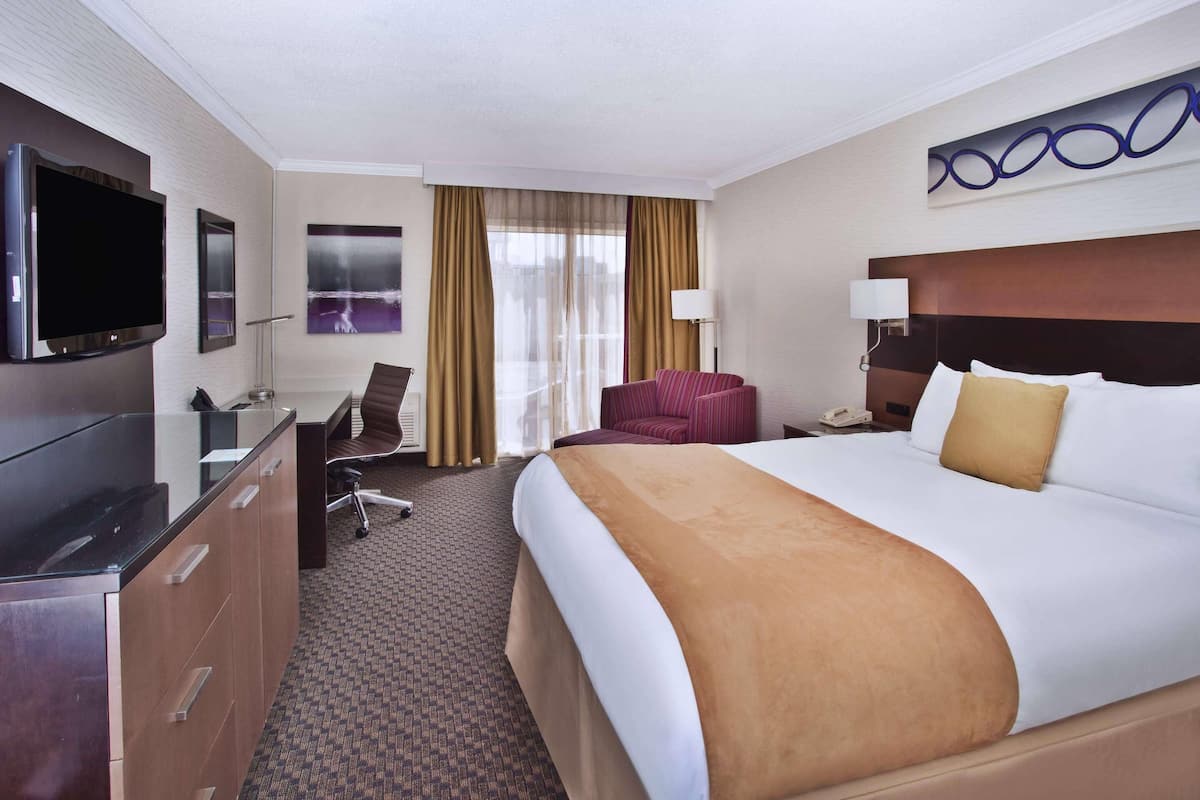 A hotel room with a large bed, a desk with a flat-screen TV, a chair, a desk lamp, and a large window with curtains.