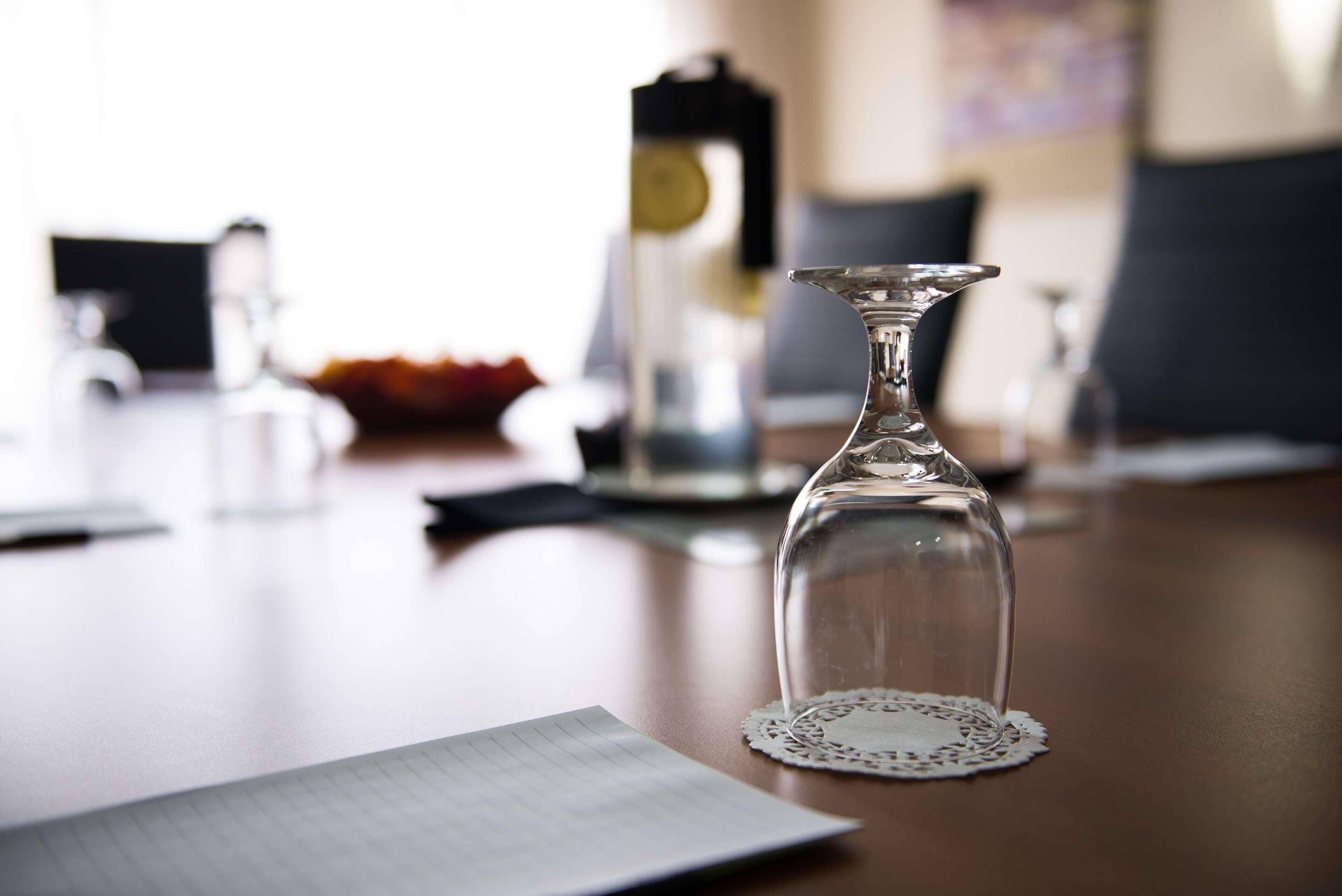 A clear glass decanter on a table with a lace doily, a notebook, and a blurred background with a lamp and a vase.