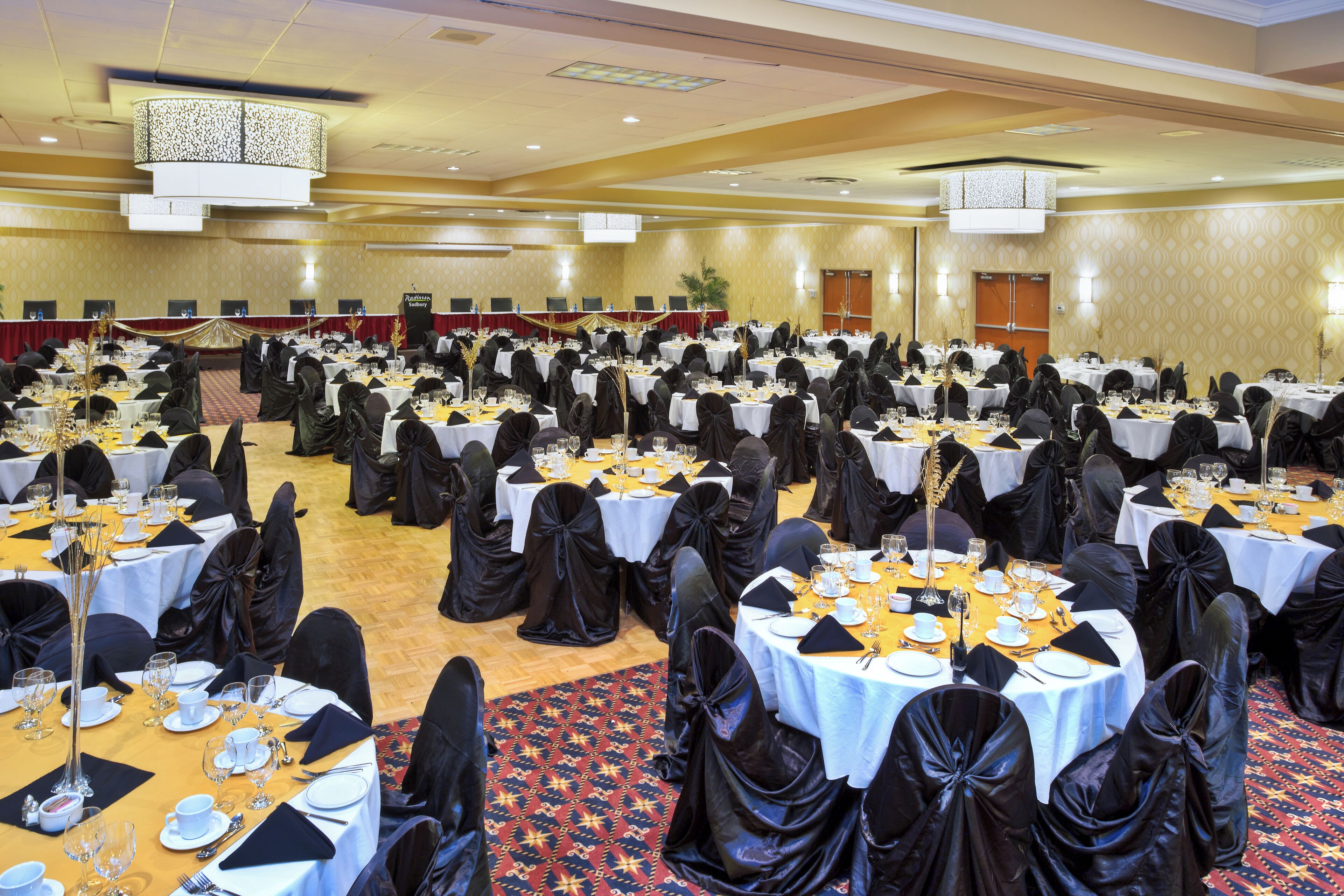 Un salón de banquetes con mesas redondas dispuestas para comer, fundas negras en las sillas y una alfombra estampada.