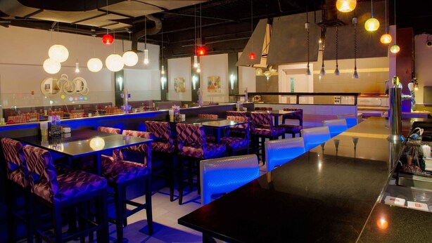 A restaurant with booths and tables, hanging pendant lights, and a counter with a blue-lit bar area. - Radisson Hotel Sudbury (Sudbury)