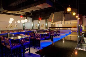 Un restaurante con cabinas y mesas, lámparas colgantes y una barra iluminada en azul.