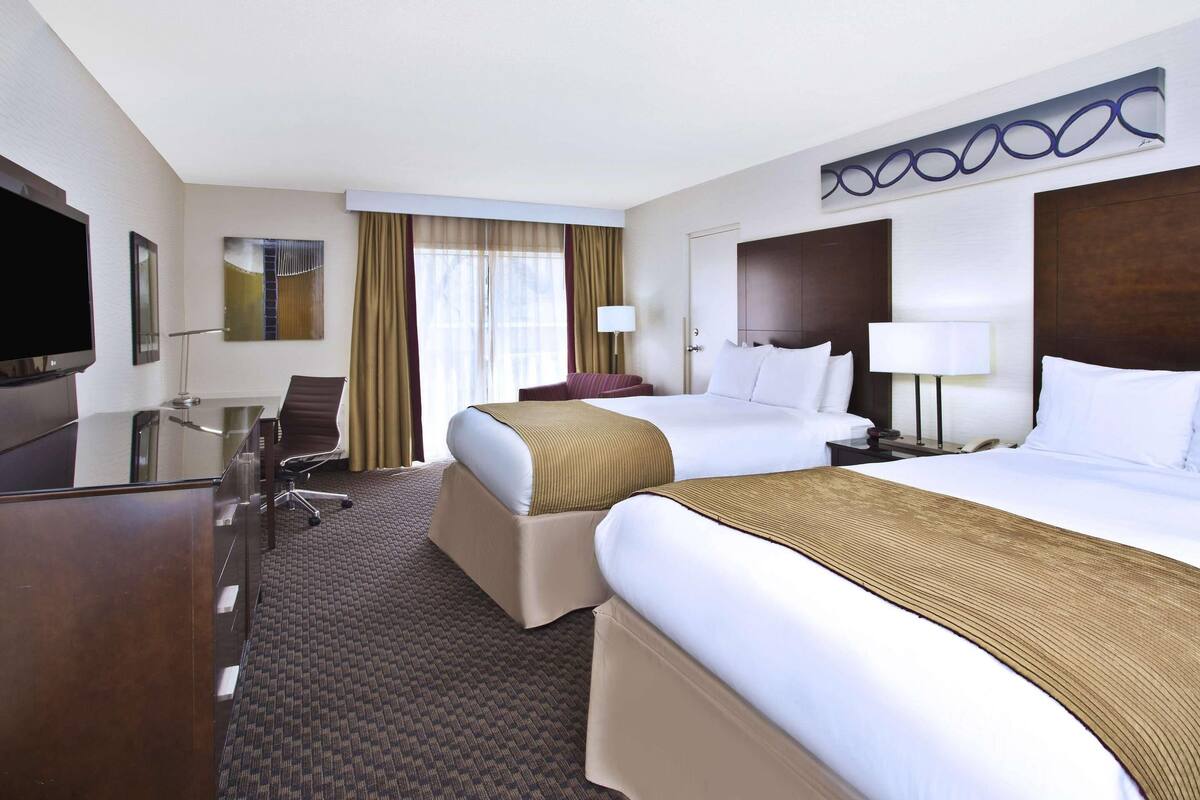 A hotel room with two beds, a desk, a TV, and a large window with curtains.