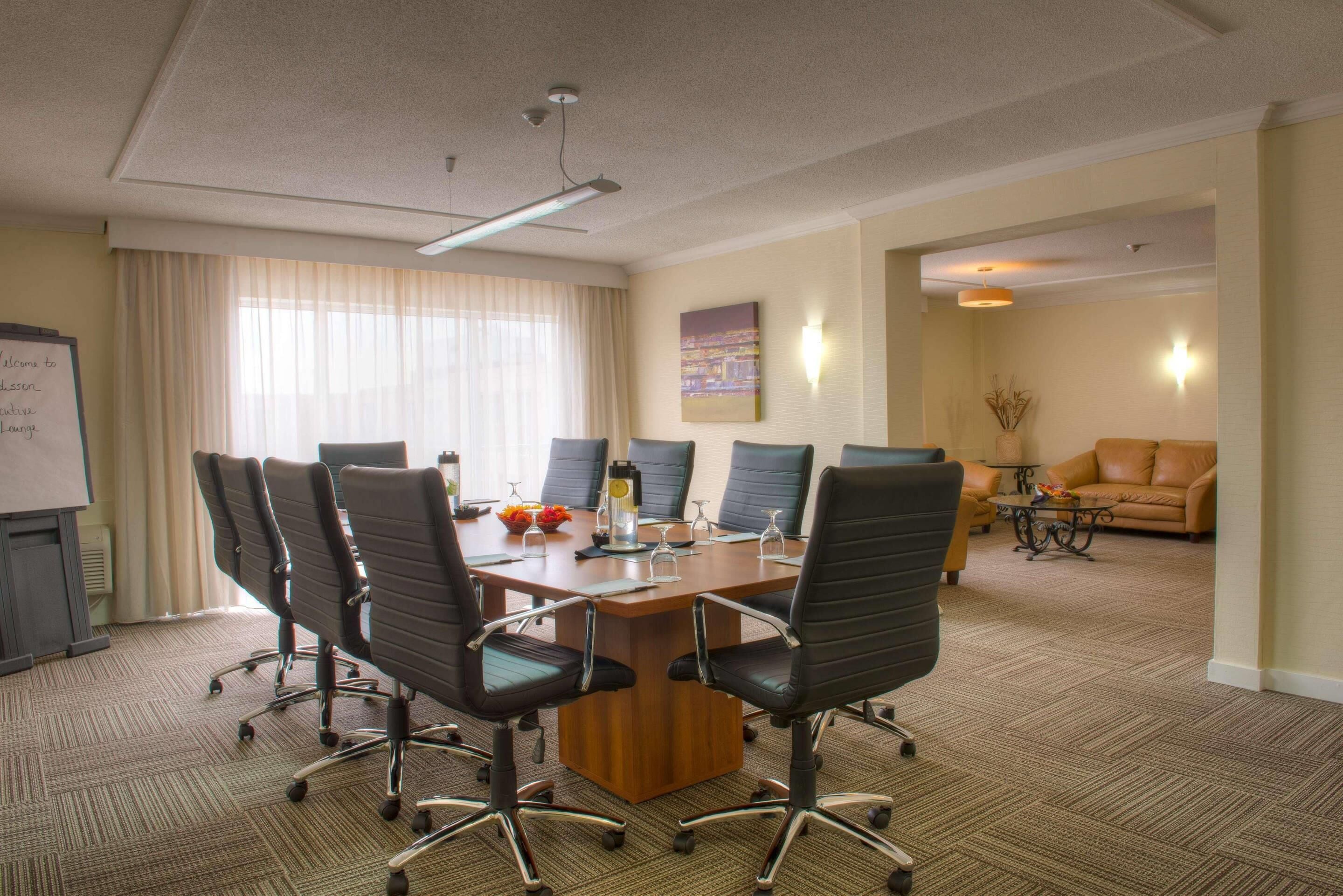 A conference room with a large table, six chairs, a whiteboard, and a sofa in the background.
