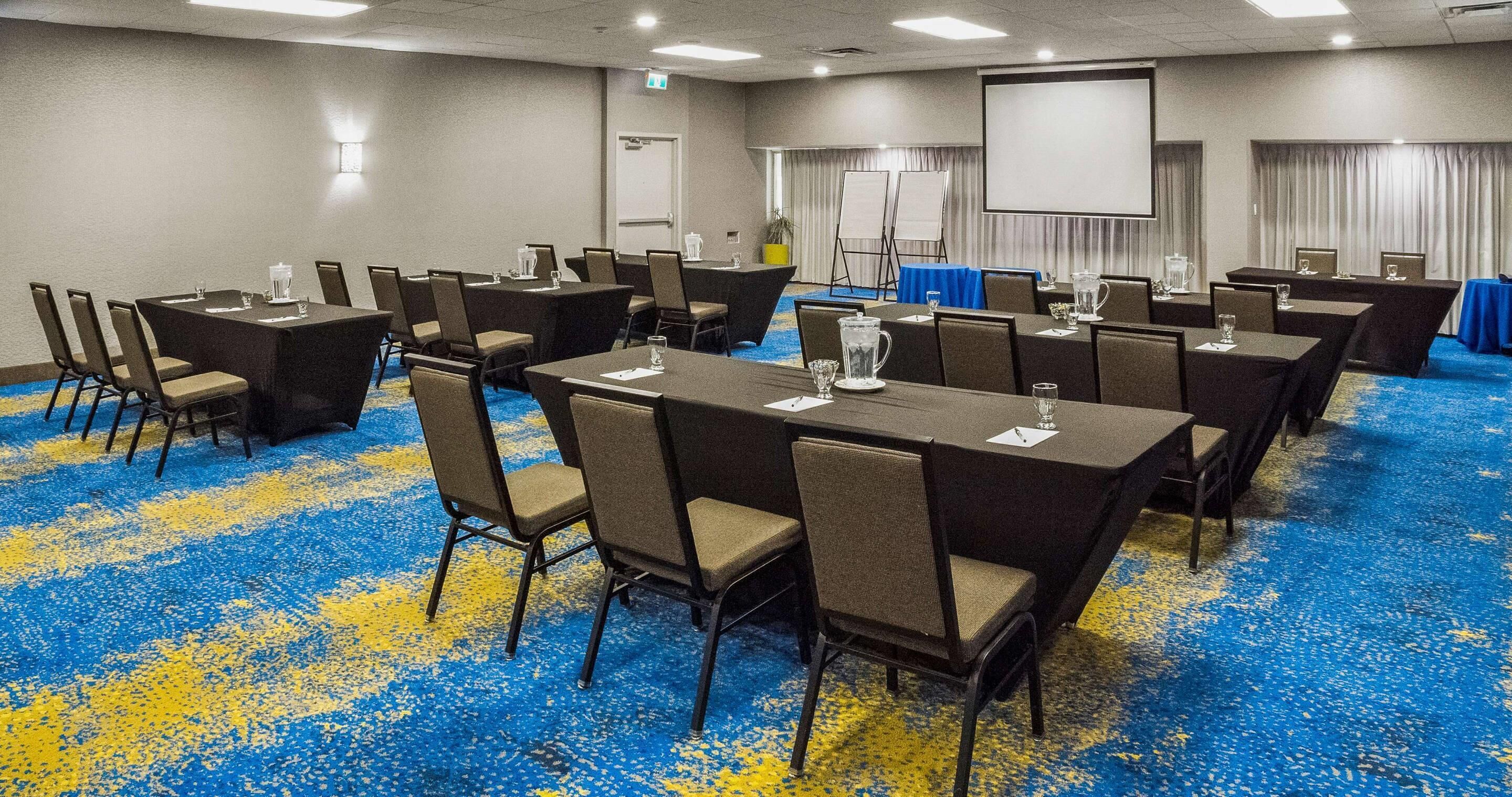 A conference room with multiple tables and chairs, a projector screen, and a carpeted floor.