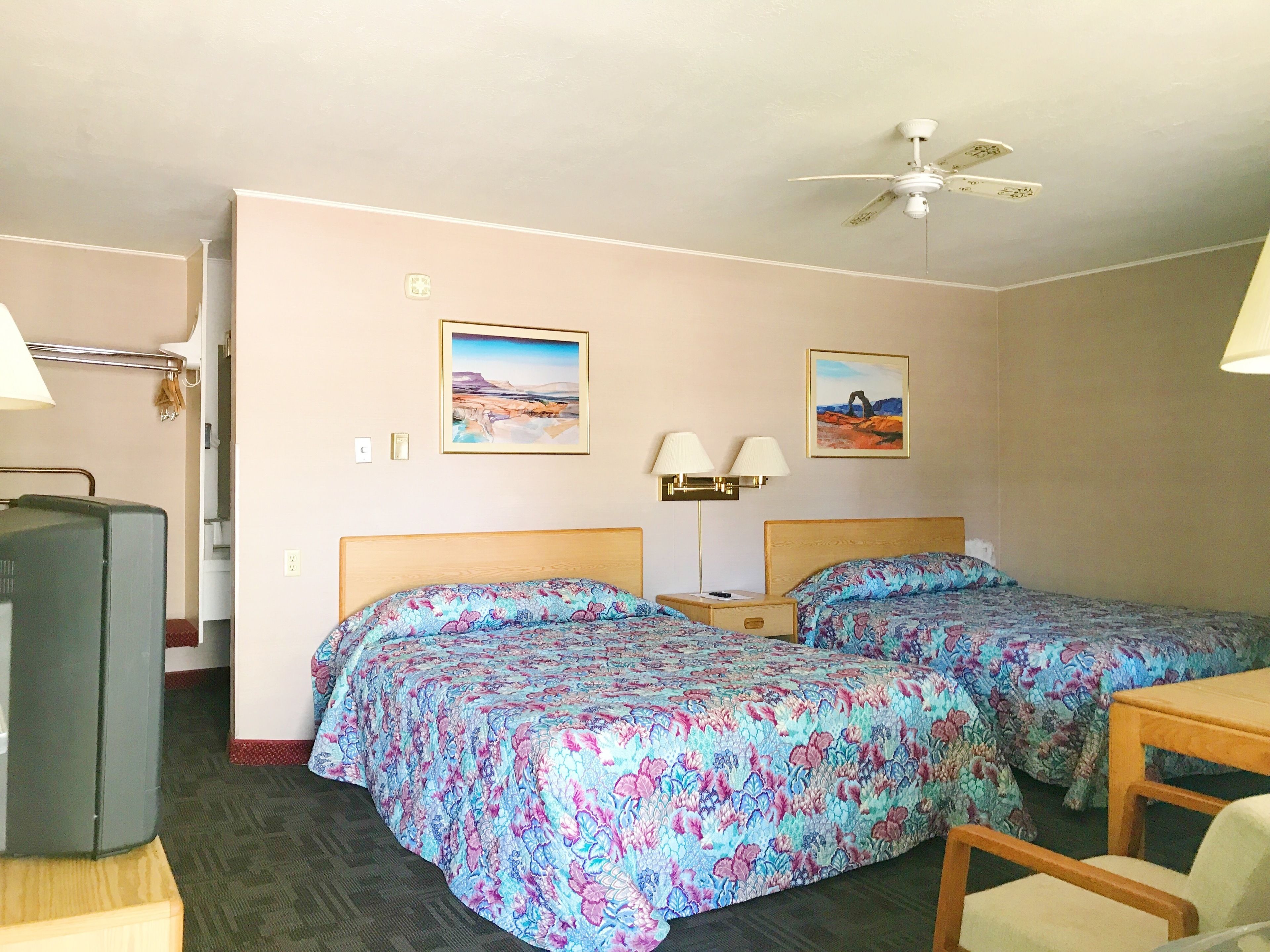 A hotel room with two beds, a desk, a chair, a television, and a ceiling fan.