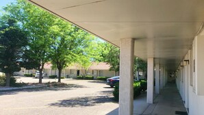 Un pasillo cubierto con columnas, una zona de estacionamiento con automóviles y árboles.