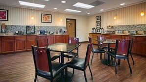 A hotel dining area with a long table and chairs, wooden cabinets, and a kitchen area with appliances. - Best Western Plus Rama Inn & Suites (La Grande)