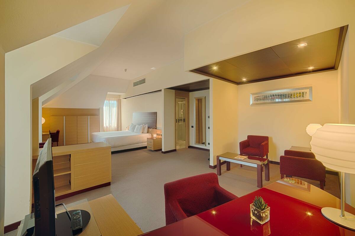 A modern hotel room with a bed, a desk, a red armchair, and a TV.