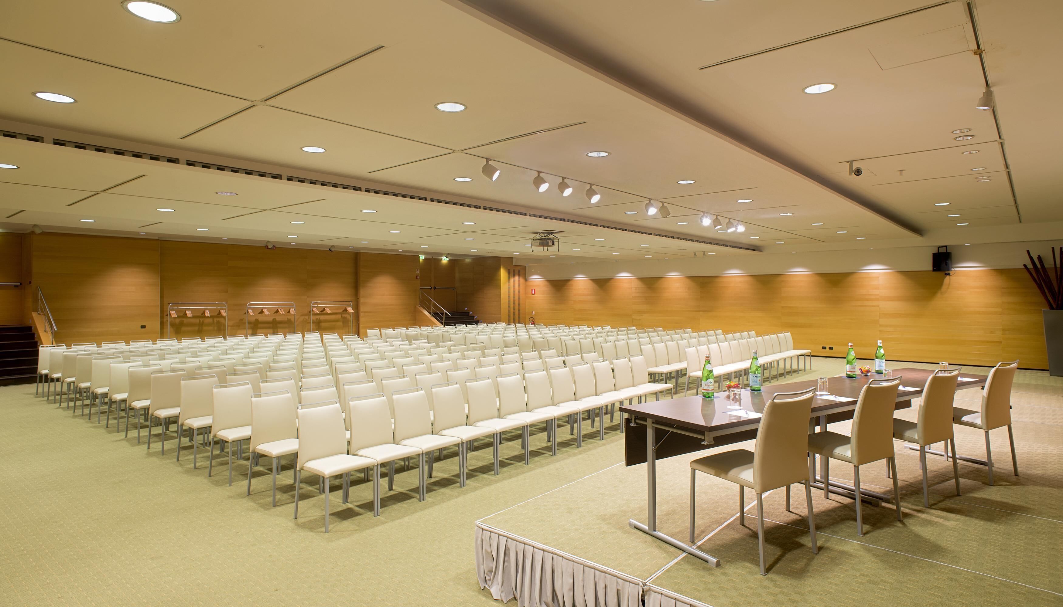 A conference room with rows of white chairs, a stage with a podium, and a table with chairs and bottles.