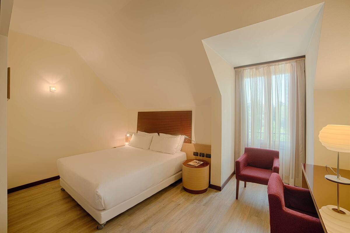 A hotel room with a large bed, a wooden headboard, a small round table, a red armchair, and a window with curtains.