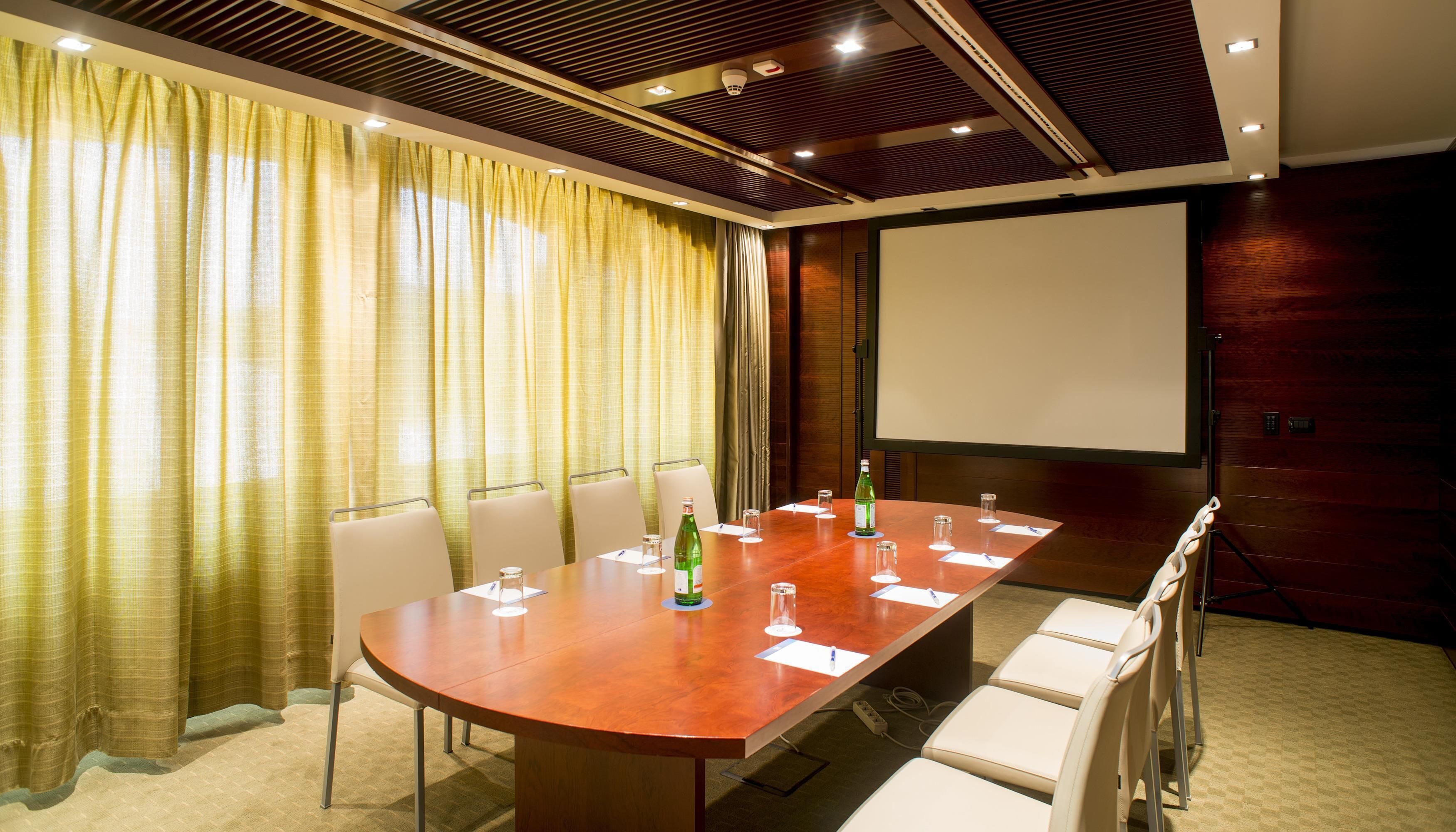 Ein Konferenzraum mit langem Tisch, Stühlen, einer Projektionsleinwand und einem großen Fenster mit Vorhängen.