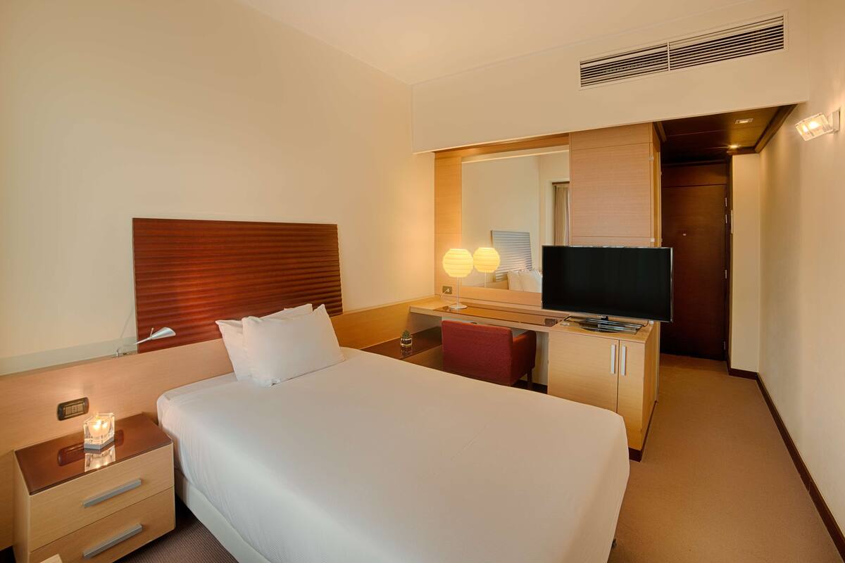 A hotel room with a large bed, a desk with a television, a mirror, and a red chair.