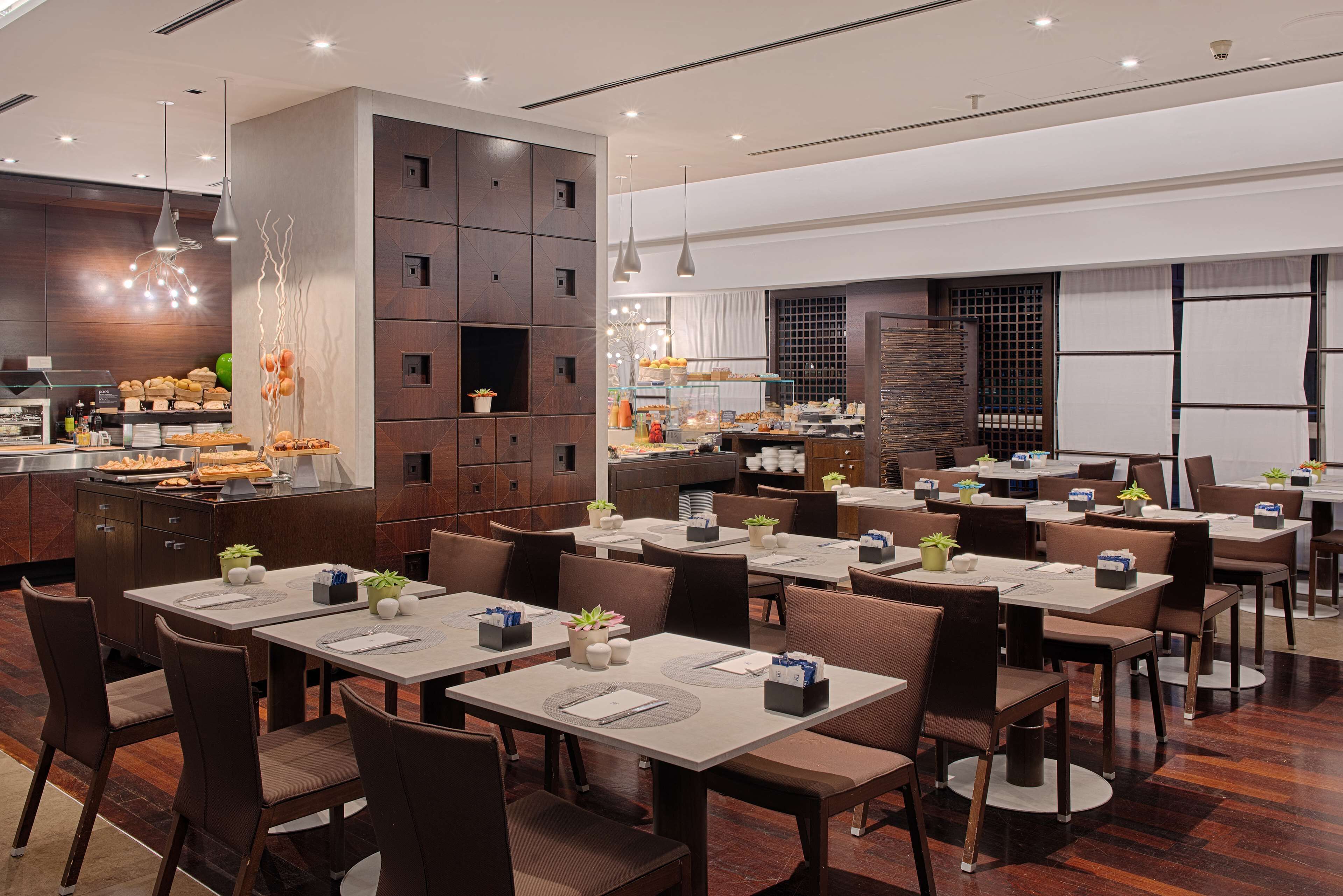 Un restaurante moderno con varias mesas y sillas, una zona de cocina con exhibición de alimentos y plantas decorativas.