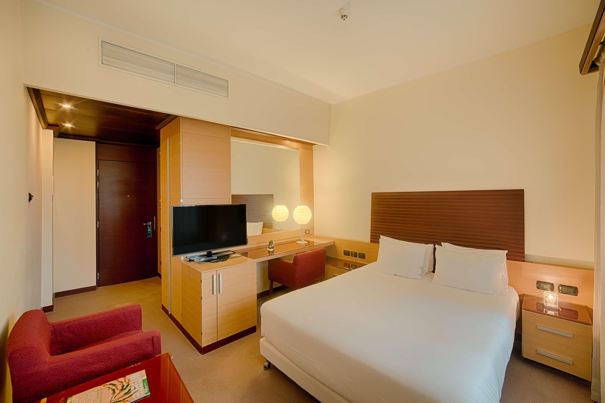 A hotel room with a bed, a desk with a TV, a red chair, a wooden nightstand, and a mirror.