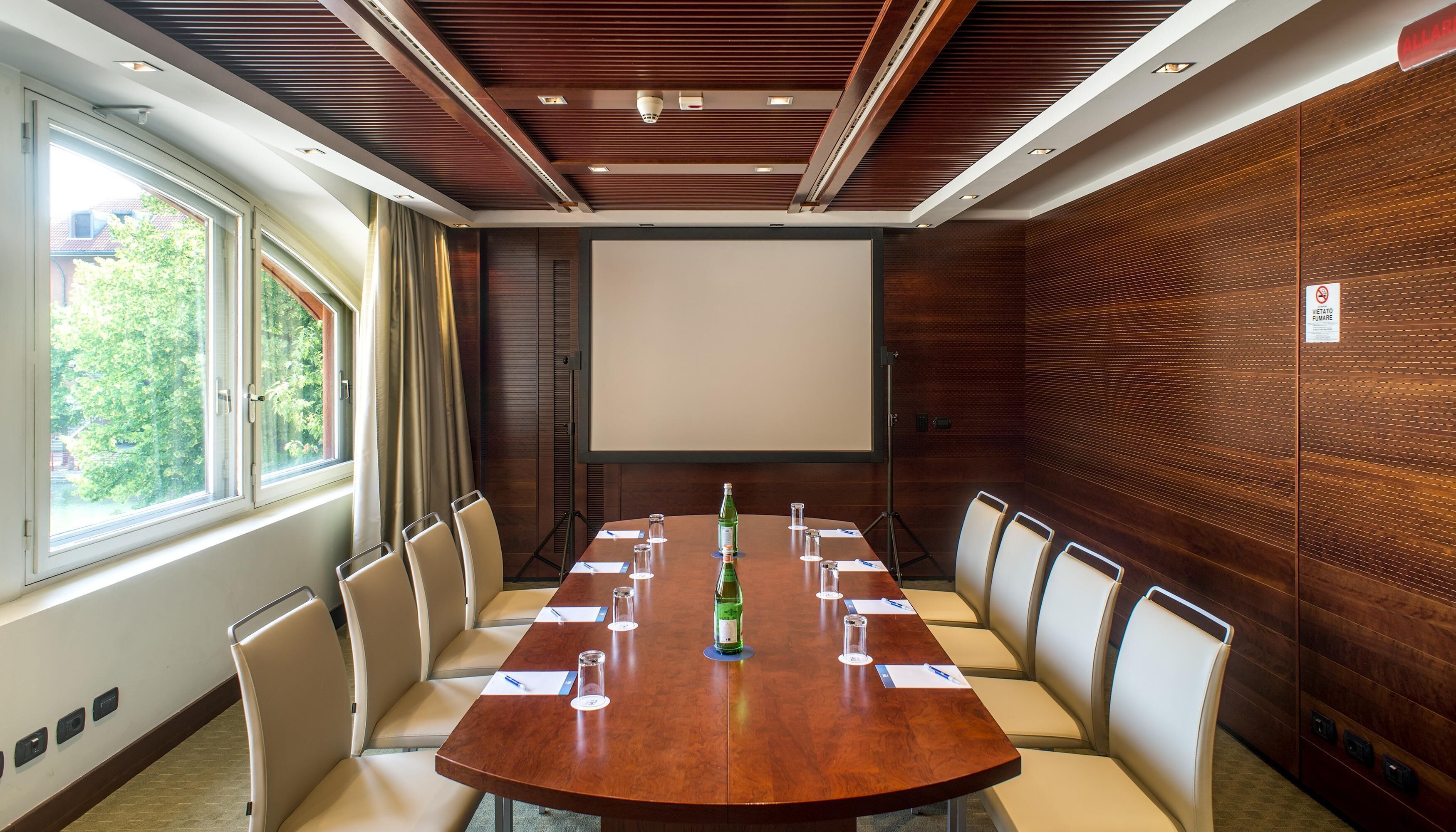 Un salón de conferencias con una larga mesa, sillas, una pantalla de proyector y una botella de agua.