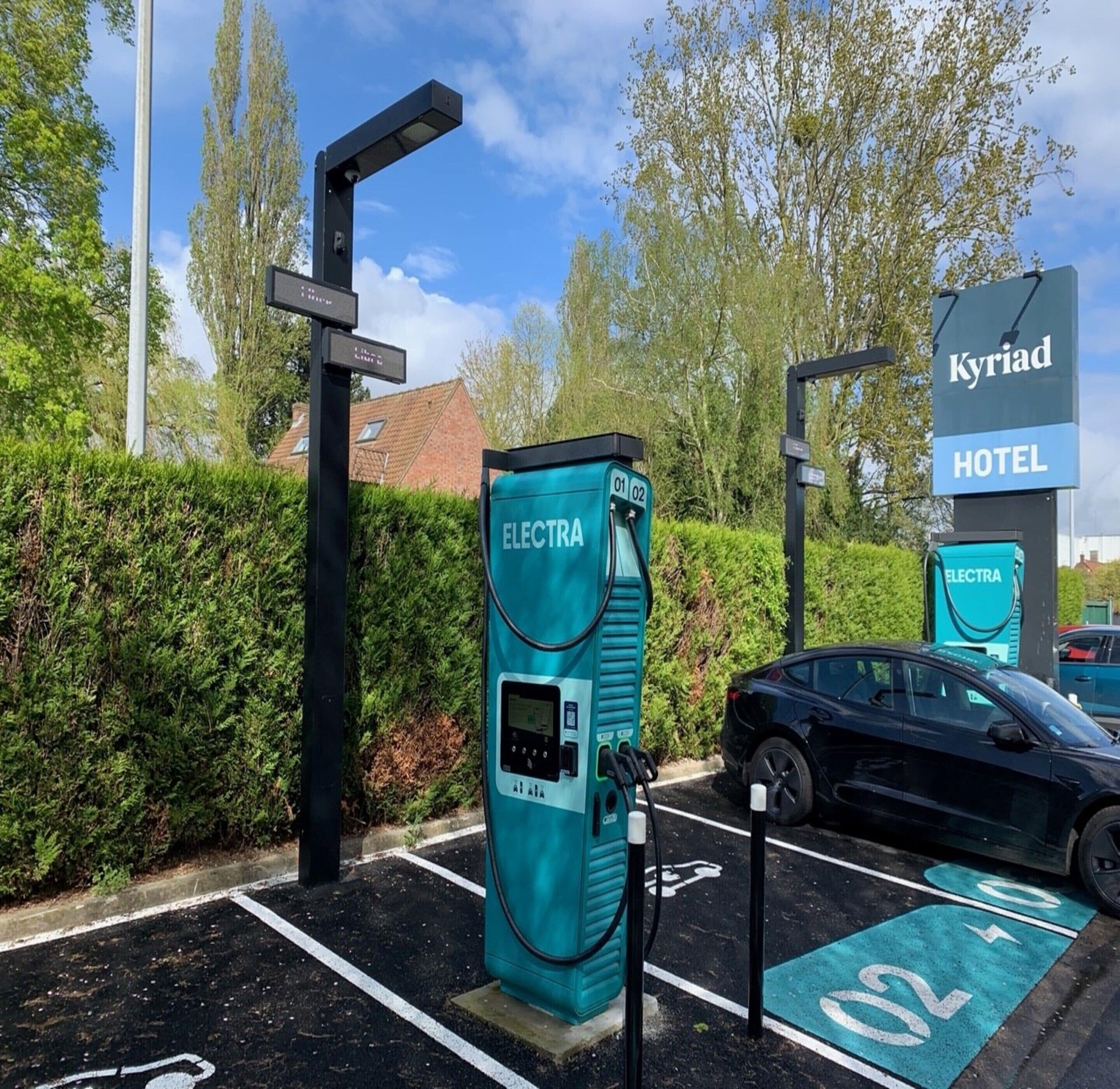 Una estación de carga para vehículos eléctricos, con un letrero del Hotel Kyriad al fondo.