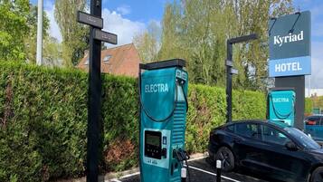 Una estación de carga para vehículos eléctricos, con un letrero del Hotel Kyriad al fondo.