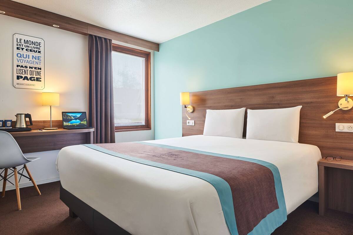 A hotel room with a large bed, a desk with a computer and lamp, a window with curtains, and a motivational quote on the wall.