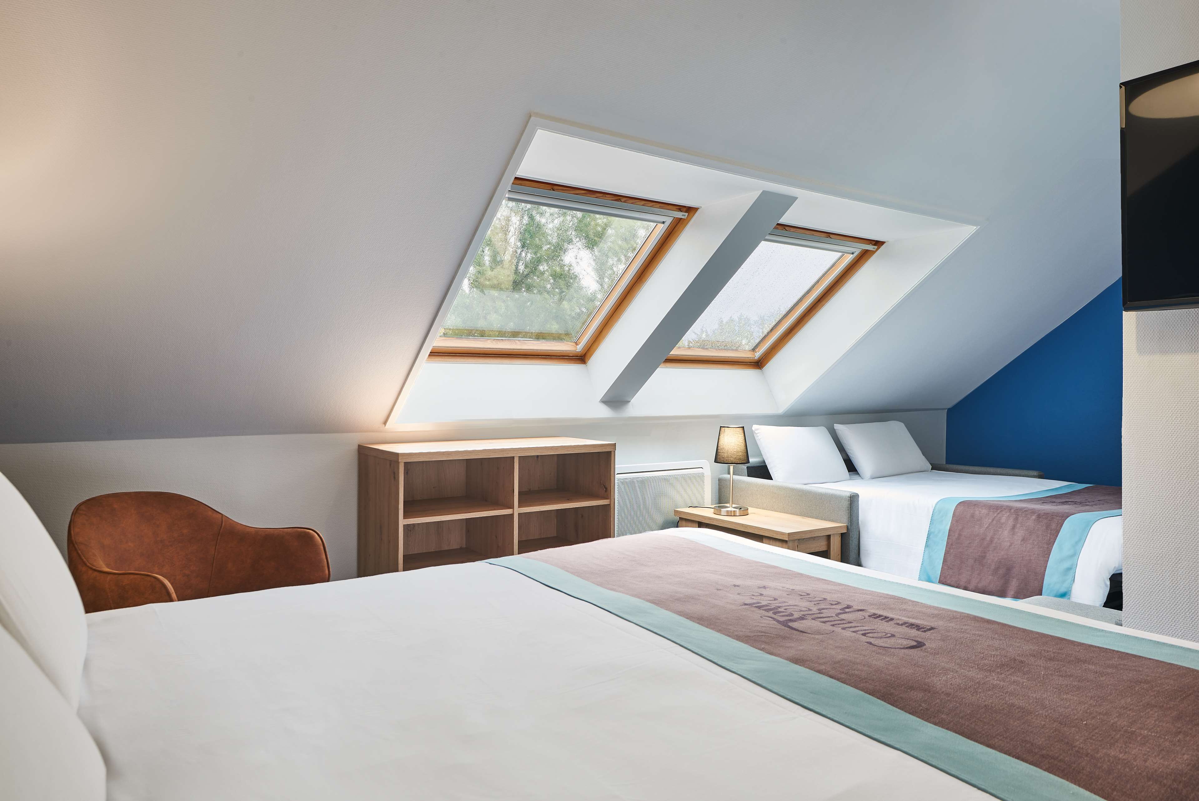 A bedroom with a sloped ceiling, two skylights, a bed, a chair, a bedside table, and a lamp.