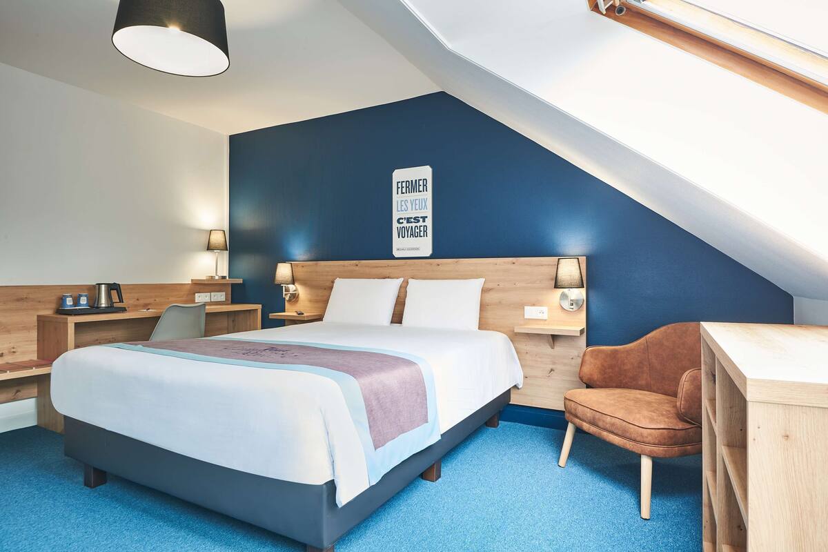 A modern hotel room with a large bed, a desk, and a chair. There is a sign on the wall that says "FERMER LES YEUX C'EST VOYAGER".