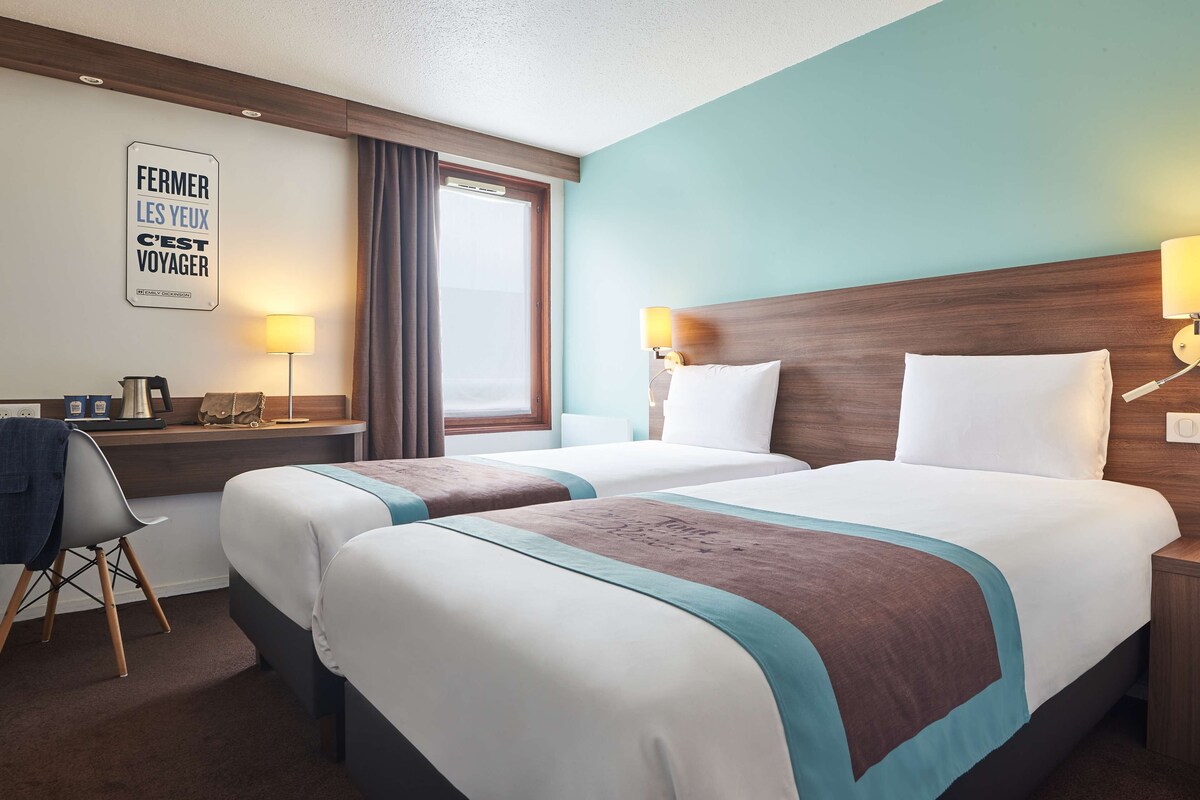 A hotel room with two beds, a desk with a lamp, a chair, and a window with curtains.