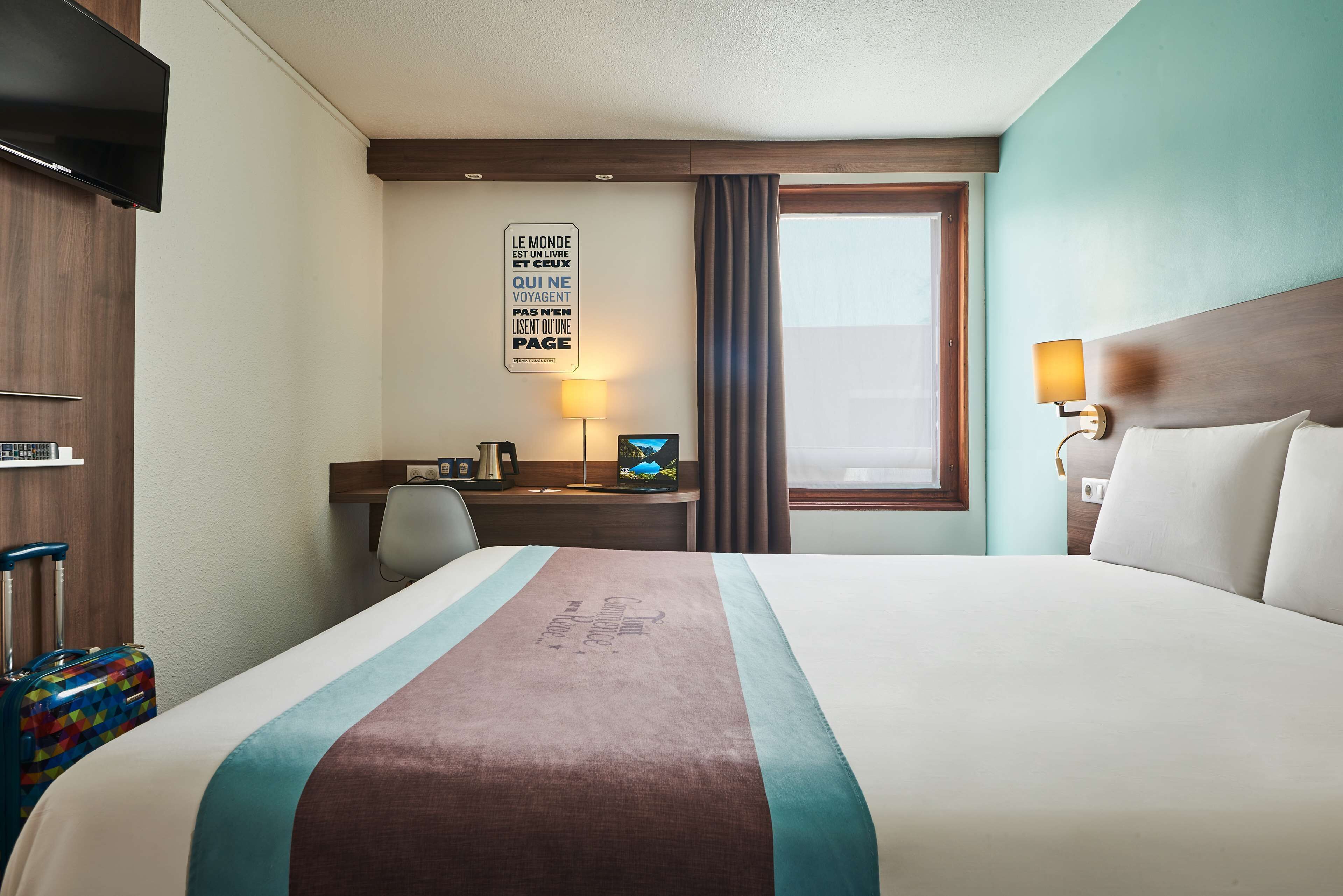 A hotel room with a large bed, a desk with a computer, a TV, and a window with curtains.