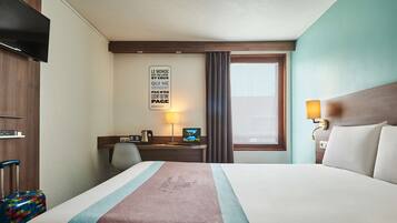 A hotel room with a large bed, a desk with a computer, a TV, and a window with curtains.
