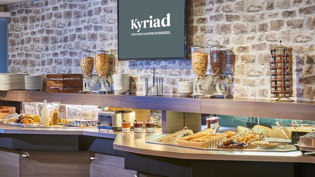 Zona de desayuno tipo buffet con pared de piedra, un televisor mostrando 'Kyriad Hotel' y diversas opciones de comida y bebida.