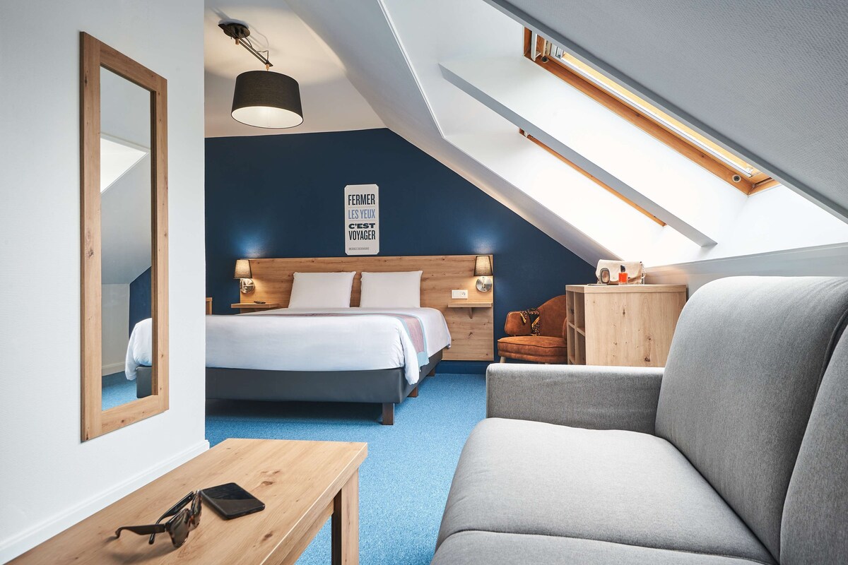 A modern hotel room with a large bed, a sofa, a wooden nightstand, and a ceiling skylight.