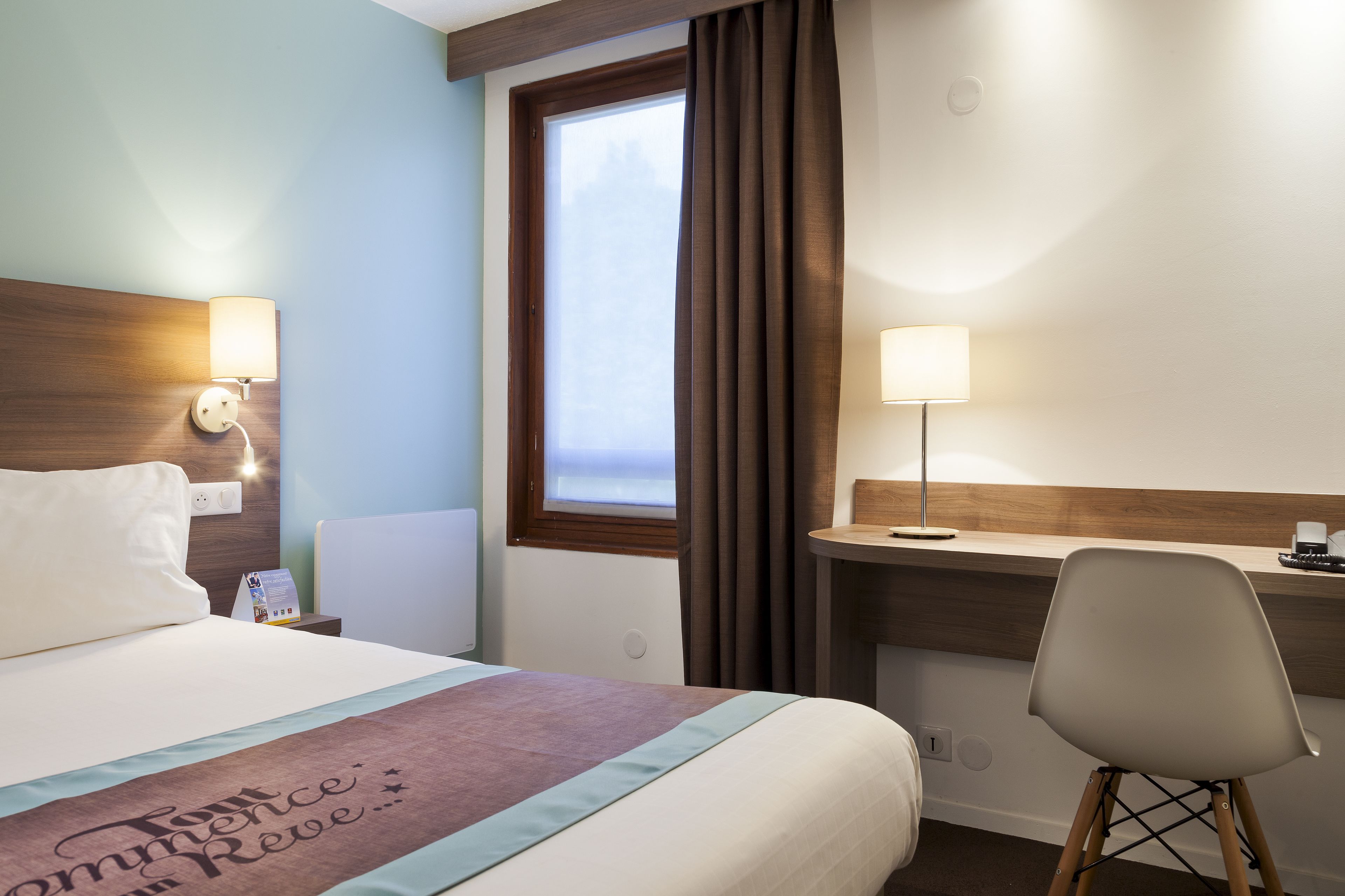 Une chambre d’hôtel comprenant un lit, une table de chevet, une lampe, un bureau, une chaise et une fenêtre avec des rideaux.