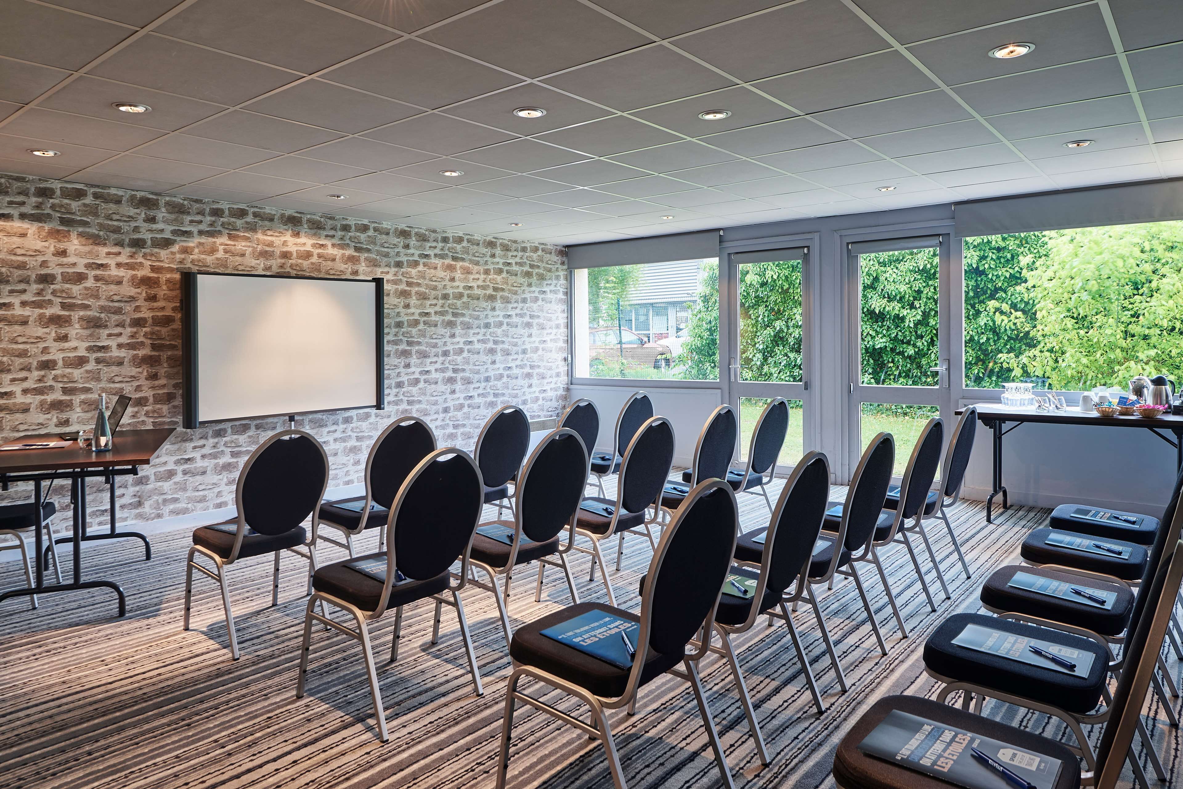 A conference room with a brick wall, a projector screen, and a large window with a view of greenery.