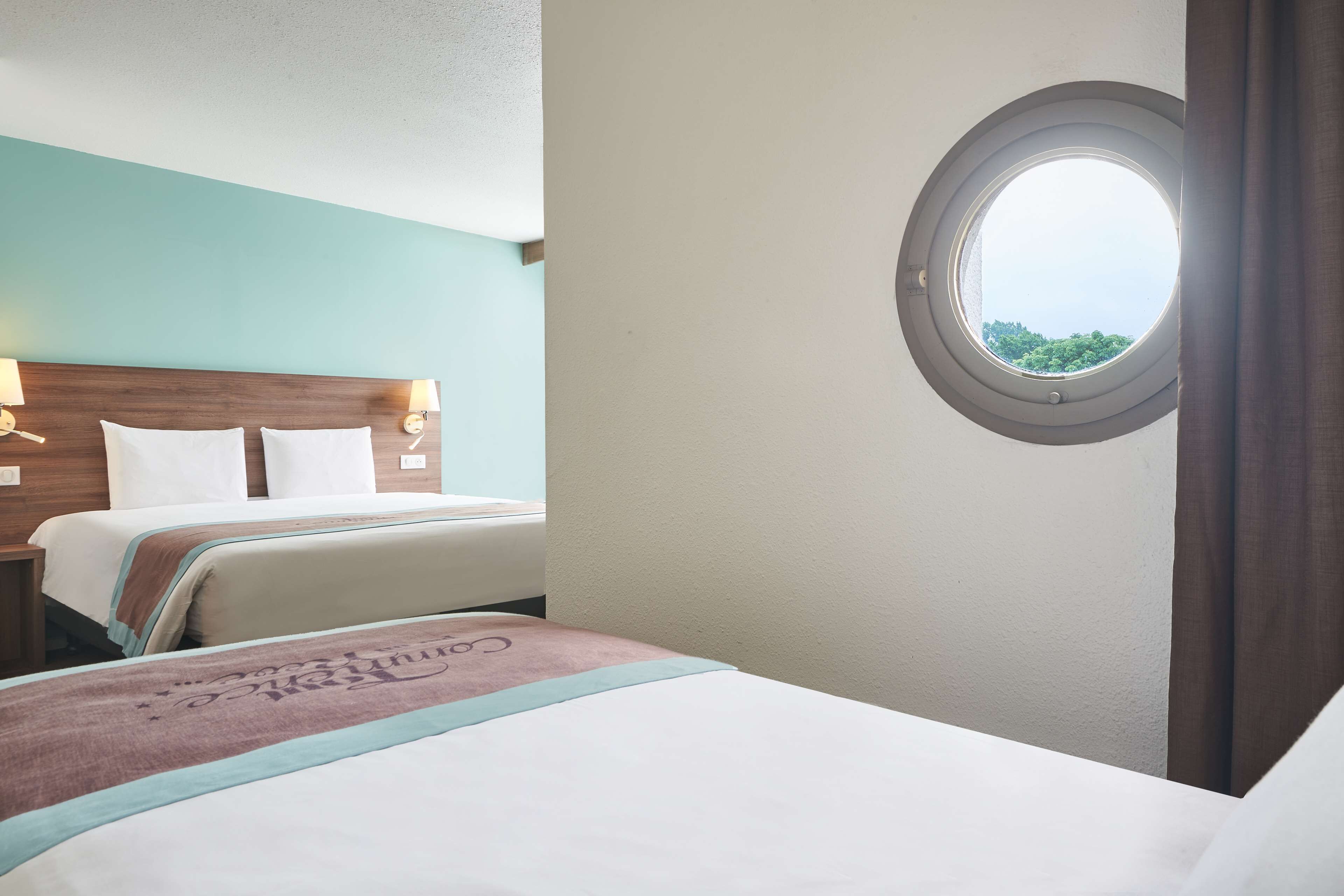 A hotel room with two beds, a round window, and a view of greenery outside.