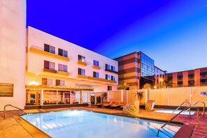 Zona de piscina de hotel con cielo despejado, una piscina, sillas de descanso y un edificio con varios pisos y balcones.