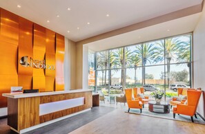 Zona de recepción de un hotel con un mostrador de madera, asientos anaranjados y grandes ventanales con vistas a palmeras y vehículos.