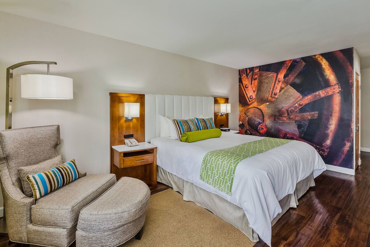 A hotel room with a bed, a chair, a nightstand, a lamp, and a large painting on the wall.