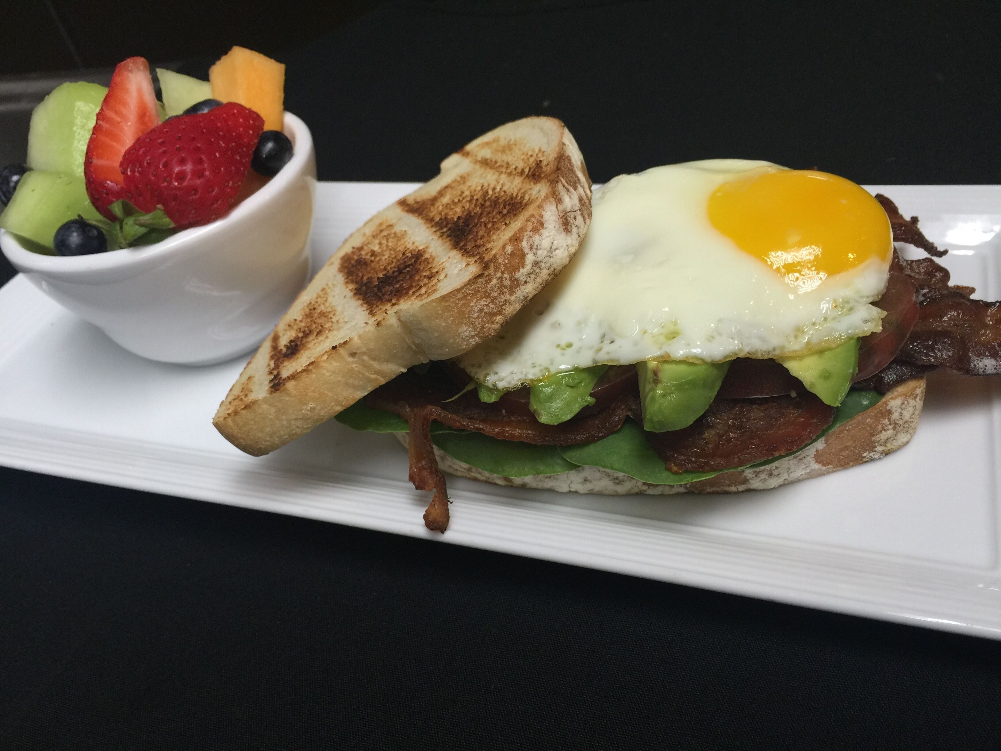 Un sándwich de desayuno con un huevo frito, tocino, aguacate y lechuga sobre un plato blanco, acompañado de un tazón de fruta.