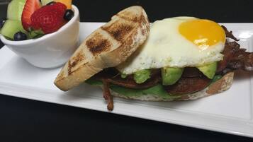 Un sándwich de desayuno con un huevo frito, tocino, aguacate y lechuga sobre un plato blanco, acompañado de un tazón de fruta.