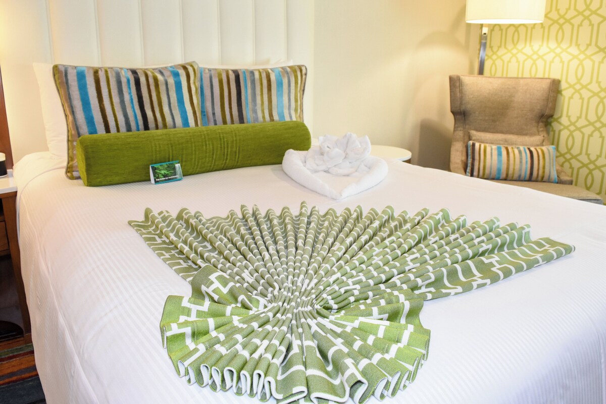 Un lit bien fait, recouvert d’une couvre-lit à motifs verts et blancs, avec un oreiller rayé et une chaise agrémentée d’un coussin rayé.