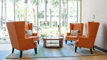 A seating area with two orange armchairs and a glass coffee table, a patterned rug, and a view of palm trees and a building through large windows.