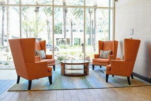 A seating area with two orange armchairs and a glass coffee table, a patterned rug, and a view of palm trees and a building through large windows. - Hotel Indigo Anaheim by IHG (Anaheim)