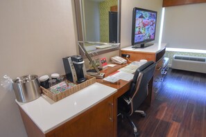 Una camera d'albergo con una scrivania in legno, un computer, una macchina per il caffè, un telefono e una finestra con vista sull'esterno.