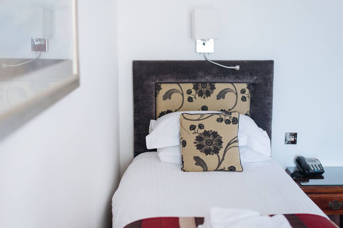 A neatly made bed with a floral-patterned headboard, a bedside phone, and a power outlet on the wall.