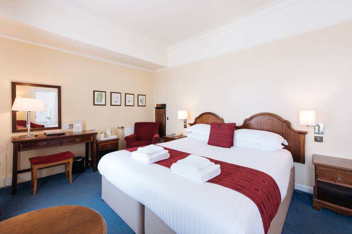 A hotel room with a large bed, a desk with a chair, a bedside table, and a lamp.