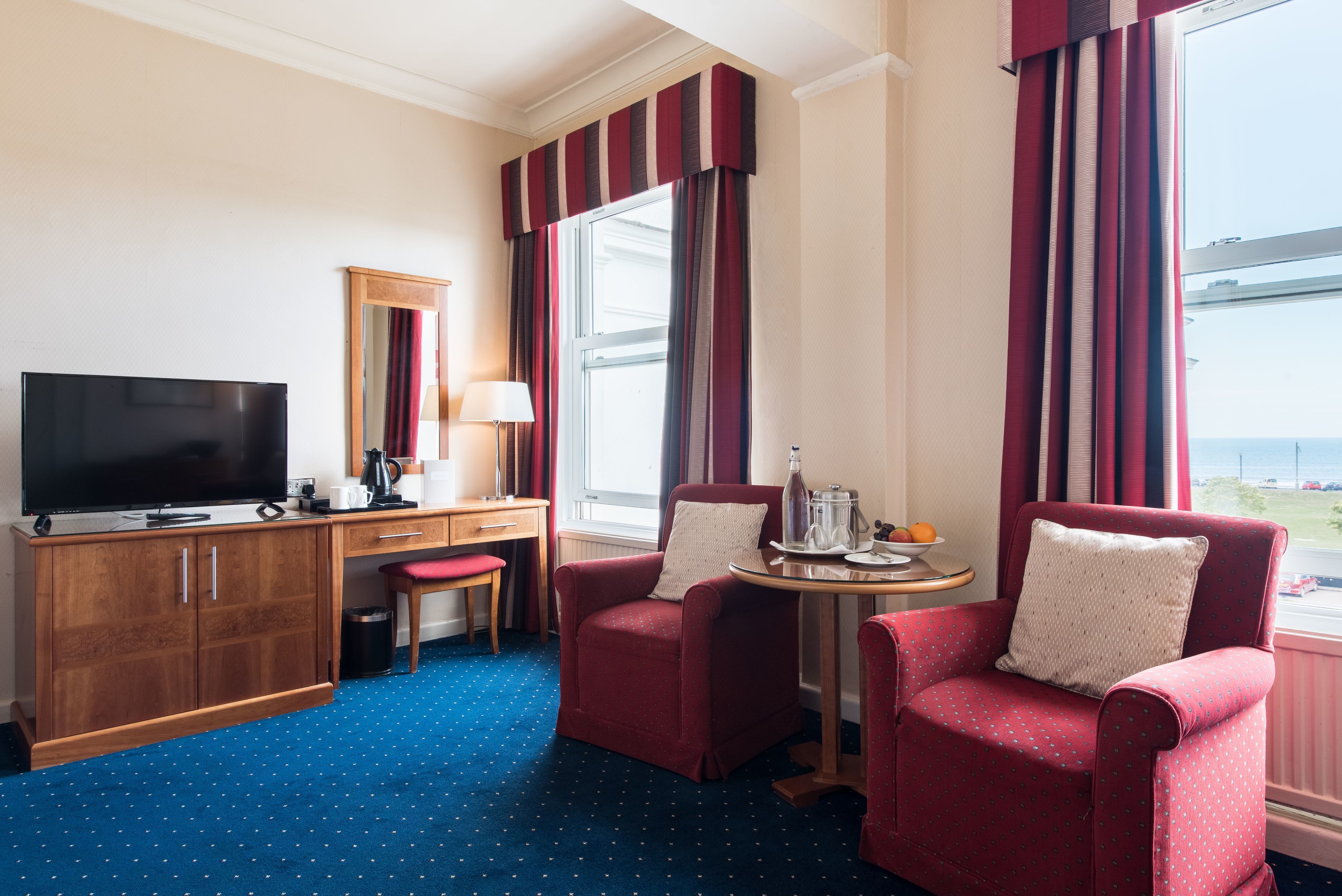 Habitación de hotel con televisión, escritorio, dos sillones, una mesita con bandeja y ventana con cortinas.